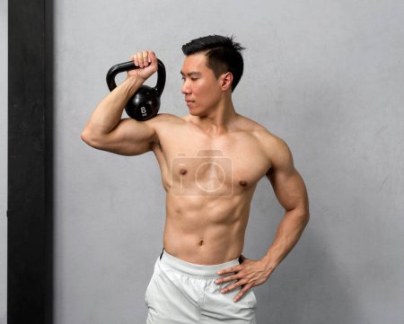 Foto de Un hombre asiático tonificado y sin camisa flexiona sus músculos, el brillo del sudor que refleja su duro trabajo, mientras hábilmente posa con una fuerte pesa en la mano. - Imagen libre de derechos