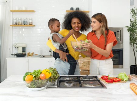 Foto de Dos jóvenes lesbianas diversas pasan tiempo en una cocina en casa, compartiendo armoniosamente las tareas de preparación de alimentos mientras alimentan simultáneamente a un bebé. - Imagen libre de derechos
