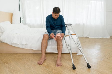 Un hombre con una rodilla rota sentado en su cama. Se apoya en muletas, señalando la recuperación de la lesión. Su dormitorio sirve como telón de fondo, teñido con toques de impotencia pero duradera determinación.