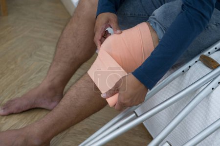 Ein Mann sitzt mit gebrochenem Knie auf seinem Bett. Er stützt sich auf Krücken, was die Genesung von Verletzungen signalisiert. Sein Schlafzimmer dient als Kulisse, gefärbt mit Andeutungen von Hilflosigkeit und doch anhaltender Entschlossenheit.