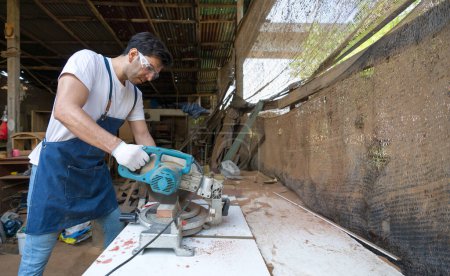 Photo for Young carpentry using a power miter saw to cut a piece of wood. Atmosphere in a workshop or outdoor shed based on the rustic environment and the presence of tools and construction materials. - Royalty Free Image