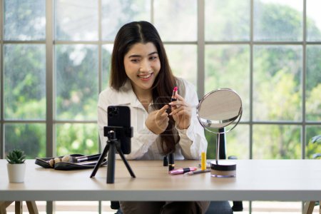 Joven mujer asiática está aplicando lápiz labial, la grabación de un tutorial de maquillaje con un teléfono inteligente y espejo.