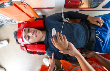 A person is lying on a stretcher with a head immobilizer and is monitored by medical personnel in orange uniform inside an ambulance. Top view