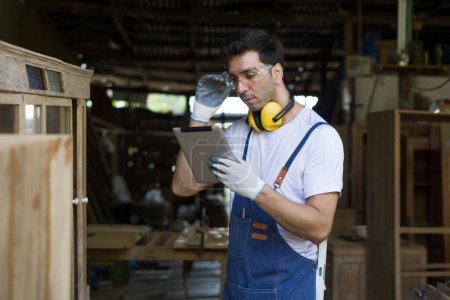 Foto de Un carpintero o carpintero sosteniendo una tableta, revisando el plan de diseño o la instrucción relacionada con la carpintería. Uso de gafas de seguridad, audífonos y guantes en un taller de carpintería. - Imagen libre de derechos