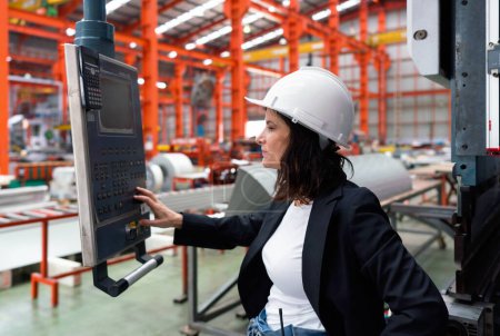 A woman in black suit and a hardhat is carefully observing a machinery monitor in a bustling factory.