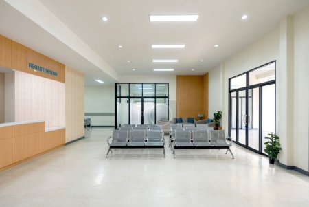 Waiting area in a hospital. Patient or visitor can check in for appointment or service. There are rows of connected grey seating for individual waiting. Glass door on the right lead to emergency area.