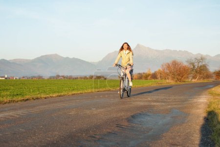 Foto de Mujer joven con chaqueta monta una bicicleta sobre el camino del campo, sol de la tarde brilla en el pico Krivan monte - Símbolo eslovaco - fondo - Imagen libre de derechos