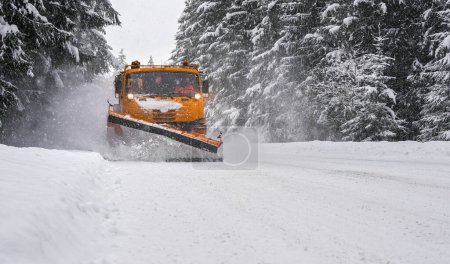 Foto de Camión de arado de mantenimiento naranja en carretera forestal después de tormenta de nieve ventisca. Las carreteras se vuelven peligrosas durante el invierno el conductor se enfrenta borrosa irreconocible . - Imagen libre de derechos