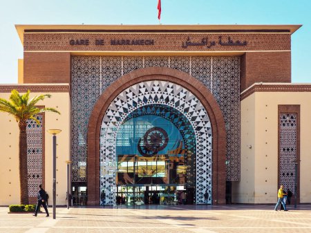 Foto de Marrakech, Marruecos - 02 de enero de 2020: Adornos que decoran las paredes de la estación de tren de Marrakech, el sol brilla en el pavimento que conduce a la gran entrada de vidrio, palma al lado - Imagen libre de derechos