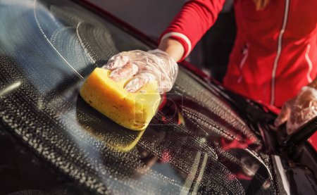 Téléchargez les photos : Jeune femme nettoyage pare-brise de sa voiture, détail gros plan à portée de main dans le gant tenant éponge jaune - en image libre de droit