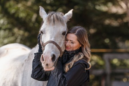 Young woman leaning to white Arabian horse, eyes closed, blurred trees background, closeup detail