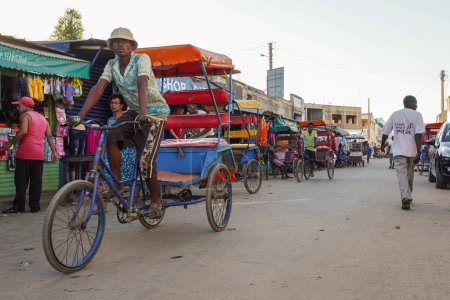 Foto de Toliara, Madagascar - 01 de mayo de 2019: Tarde en la típica calle de la ciudad por la noche, puestos de mercado al lado, gente malgache caminando, algunas bicicletas de montar, tres bicicletas taxi en primer plano - Imagen libre de derechos