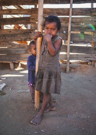 Téléchargez les photos : Ilakaka, Madagascar - 30 avril 2019 : Un enfant malgache à l'air pauvre et inconnu, debout dans la rue à côté d'une cabane en bois, portant des vêtements donnés par des touristes - en image libre de droit