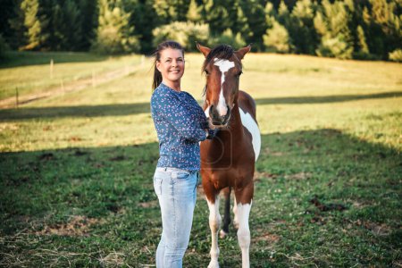 Téléchargez les photos : Femme en jean et chemise debout à côté de poulain cheval pinto brun et blanc, prairie verte derrière elle, gros plan photo - en image libre de droit