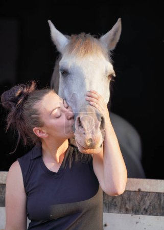 Téléchargez les photos : Jeune femme debout à côté du cheval arabe blanc reposant dans une boîte d'écuries en bois, l'embrassant les yeux fermés, le soleil brille sur eux, détail gros plan - en image libre de droit