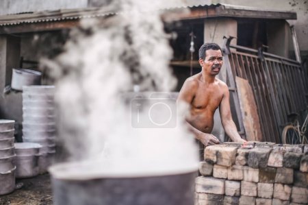 Téléchargez les photos : Ambatolampy, Madagascar - 25 avril 2019 : Usine de traitement de l'aluminium, boîtes fondent dans des pots avec de l'eau bouillante sur le feu à l'extérieur, vapeur au-dessus, travailleur inconnu debout à côté du four à briques - en image libre de droit