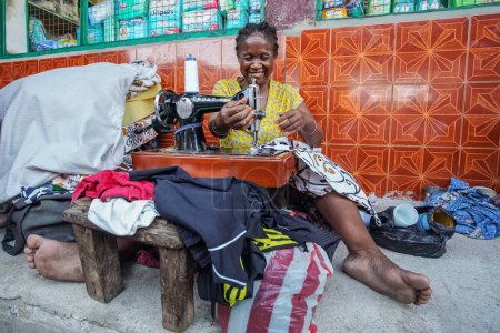 Téléchargez les photos : Toliara, Madagascar - 01 mai 2019 : Femme malgache âgée inconnue assise par terre dans la rue à côté du marché, réparant des vêtements avec des machines à coudre à manivelle - en image libre de droit