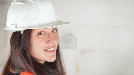 Téléchargez les photos : Jeune femme en casque blanc et gilet orange haute visibilité, longs cheveux foncés, regardant par-dessus son épaule dans la caméra, souriant. Fond flou du mur du chantier de construction, espace pour le texte côté droit - en image libre de droit