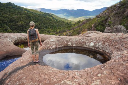 Téléchargez les photos : Jeune femme vêtue de vêtements de randonnée, debout sur des pierres près de la piscine d'eau naturelle, des arbres flous et un fond de montagne, vue derrière - paysages typiques vus lors de trek à Andringitra, Madagascar - en image libre de droit