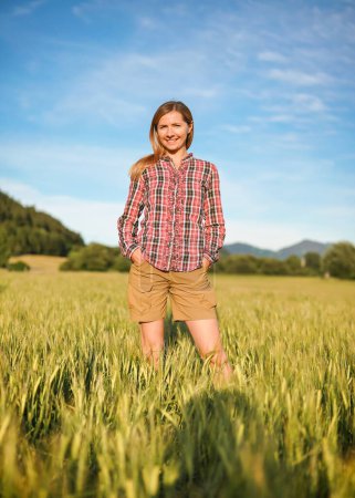 Téléchargez les photos : Jeune femme en chemise à carreaux gingham et short debout dans un champ de blé vert non mûr, souriant, ciel bleu et paysage flou avec un petit fond de forêt - en image libre de droit