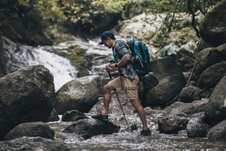 Téléchargez les photos : Les randonneurs utilisent un bâton de trekking avec des sacs à dos marchant à travers la roche et l'eau sur le ruisseau dans la forêt. concept de randonnée et d'aventure. - en image libre de droit