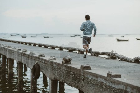 Foto de Estilo de vida saludable joven fitness hombre corriendo a la orilla del mar viejo puente. Entrenamiento al aire libre, Concepto de estilo de vida saludable. - Imagen libre de derechos
