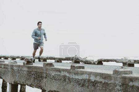 Foto de Estilo de vida saludable joven fitness hombre corriendo a la orilla del mar viejo puente. Entrenamiento al aire libre, Concepto de estilo de vida saludable. - Imagen libre de derechos