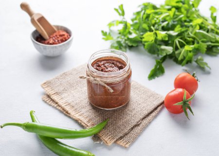 Hot sauce adjika. Homemade appetizer with pepper and tomatoes in a jar on light background with fresh herb and vegetables. The concept of Caucasian and Georgian food.