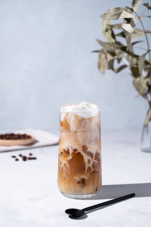 Iced coffee latte in a tall glass with milk on a light background with coffee beans, spoon and morning shadows. Summer refreshment concept. Front view.