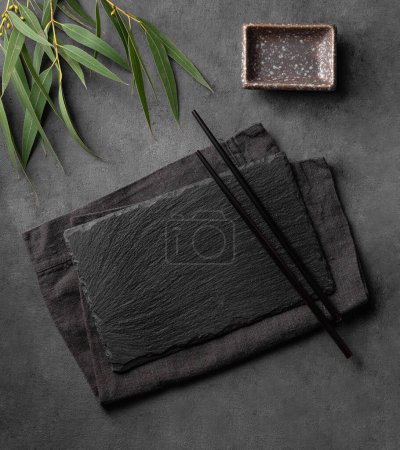 Sushi set with black chopsticks, napkin and sauce plate with green leaves on a textured dark background. Table setting concept for serving traditional Japanese food. Top view and copy space.