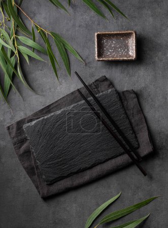 Sushi set with black chopsticks, napkin and sauce plate with green leaves on a textured dark background. Table setting concept for serving traditional Japanese food. Top view and copy space.