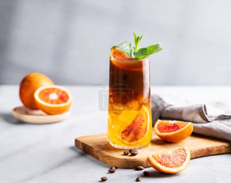 Coffee with orange juice (bumble) in a tall glass with ice and mint on a wooden board on a light background with coffee beans, fruits and morning shadows. Trendy summer Asian refreshing drink.