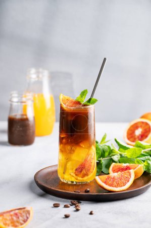 Coffee with orange juice (bumble) in a tall glass with ice and mint on a wooden plate on a blue background with coffee beans, fruits and morning shadows. Trendy summer Asian refreshing drink.