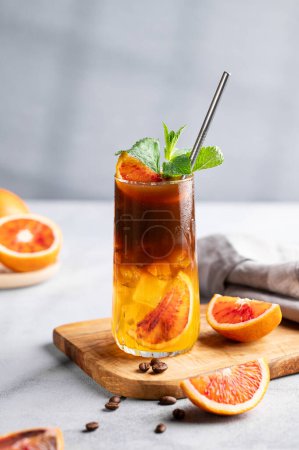 Coffee with orange juice (bumble) in a tall glass with ice and mint on a wooden board on a light background with coffee beans, fruits and morning shadows. Trendy summer Asian refreshing drink.