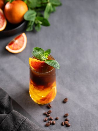 Coffee with orange juice (bumble) in a tall glass with ice and mint on a dark background with coffee beans and citrus fruits. Trendy summer Asian refreshing drink. Copy space.