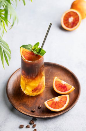 Coffee with orange juice (bumble) in a tall glass with ice and mint on a wooden plate on a blue background with coffee beans, fruits and palm leaves. Trendy summer Asian refreshing drink.