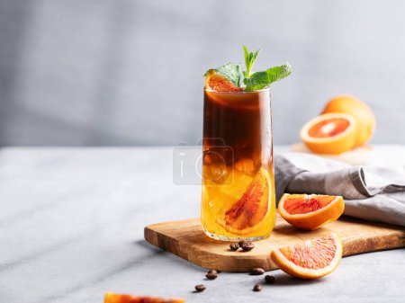 Coffee with orange juice (bumble) in a tall glass with ice and mint on a wooden board on a light background with coffee beans, fruits and morning shadows. Trendy summer Asian refreshing drink. 