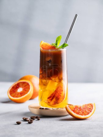 Espresso coffee with orange juice (bumble) in a tall glass with ice and mint on a blue background with coffee beans, citrus fruits and morning shadows. Trendy summer Asian refreshing drink.