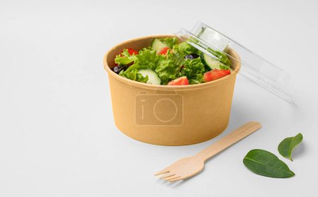 Salade verte fraîche avec tomates et concombres dans un récipient en papier avec une fourchette en bois sur un fond clair. Concept d'alimentation saine dans des plats à emporter biodégradables respectueux de l'environnement. Espace de copie.