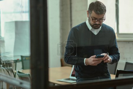 Téléchargez les photos : Homme caucasien dans des lunettes en utilisant son téléphone portable pour envoyer un message texte dans le bureau - en image libre de droit