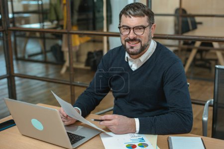 Téléchargez les photos : Happy male businessman holding papers while using laptop at his workplace - en image libre de droit