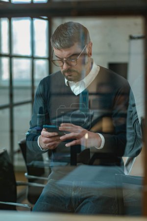 Téléchargez les photos : Homme adulte occupé tenant son smartphone pour envoyer un message tout en restant debout dans le bureau - en image libre de droit