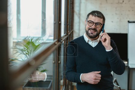 Téléchargez les photos : Smiling adult man talking on mobile phone and looking away in the office - en image libre de droit