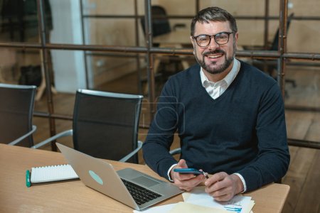 Téléchargez les photos : Homme heureux dans les lunettes tenant le téléphone portable et le stylo, travaillant avec un ordinateur portable dans le bureau moderne - en image libre de droit