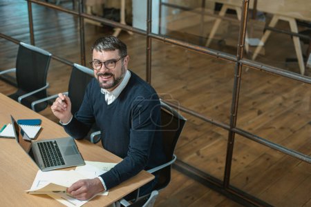 Téléchargez les photos : Top view of smiling male manager sitting at workplace while using laptop and reading documents - en image libre de droit