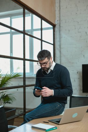 Téléchargez les photos : Concentrated man using mobile phone while typing message in the modern office - en image libre de droit