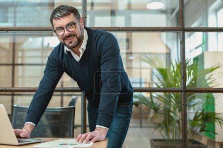 Téléchargez les photos : Happy male manager using laptop and papers while leaning on work desk - en image libre de droit