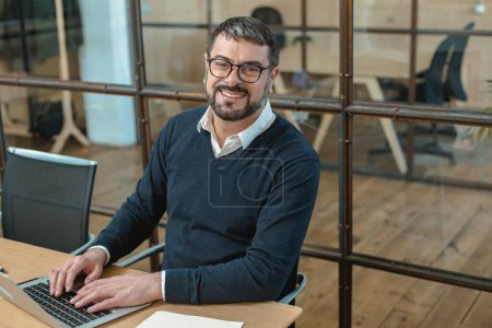 Téléchargez les photos : Happy male employee with paper documents on the desk, typing on laptop in the office - en image libre de droit