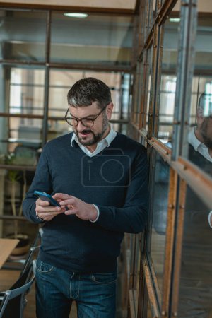 Téléchargez les photos : Happy businessman in glasses standing and holding smartphone while typing message in the office - en image libre de droit