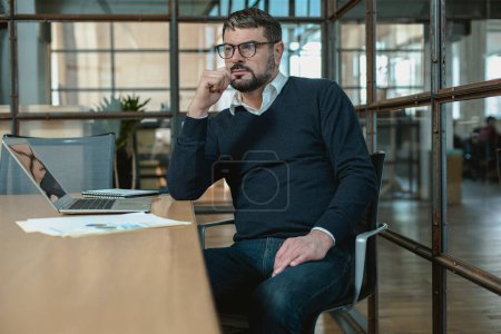 Téléchargez les photos : Barbu homme portant des lunettes de travail wih ordinateur portable et assis au bureau dans le bureau - en image libre de droit